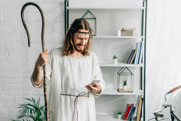 emotional Jesus in robe with wooden staff using smartphone at home - Foto, imagen