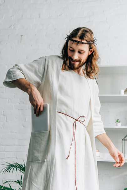 selective focus of happy Jesus in crown of thorns putting digital tablet in pocket of robe at home - Photo, image