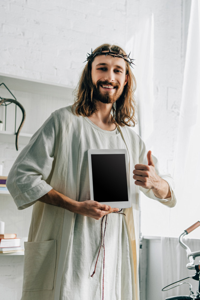 Jesus feliz na coroa de espinhos fazendo gesto polegar e mostrando tablet digital com tela em branco em casa
 - Foto, Imagem