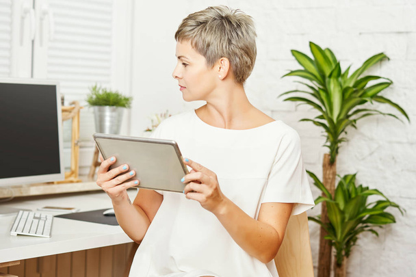 beautiful business woman in a white dress sitting at a desk and works on a tablet - Photo, Image