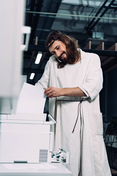 fröhlicher Jesus in Dornenkrone und Robe im Gespräch mit Smartphone und Kopierer im modernen Büro  - Foto, Bild