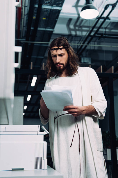 focused Jesus in crown of thorns and robe checking papers near copy machine in modern office  - Photo, Image