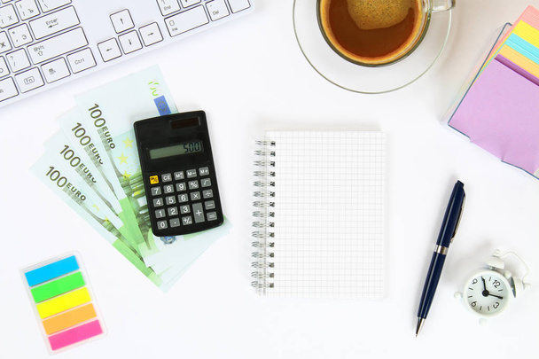 Calculator, euro banknotes, blank notebook on a white desktop next to a coffee mug and a keyboard. Top view, flat layout. Copy the space - Photo, image