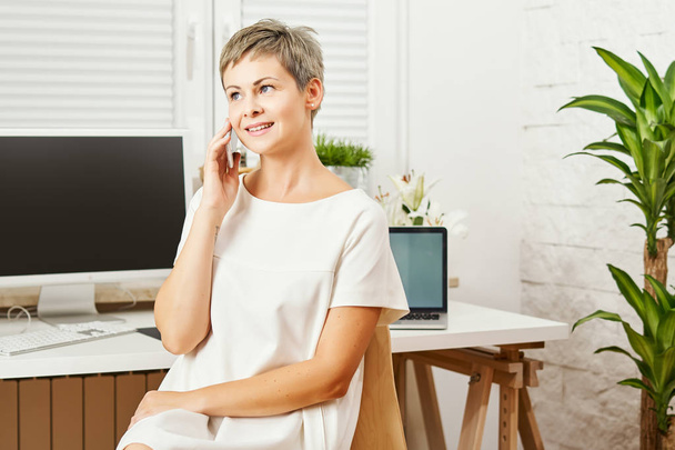 belle femme d'affaires dans une robe blanche assise à un bureau avec un téléphone à la main et travaille
 - Photo, image
