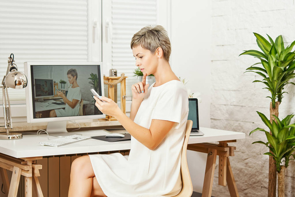 kaunis liike nainen valkoisessa mekossa istuu pöydän ääressä puhelin kädessä ja toimii
 - Valokuva, kuva