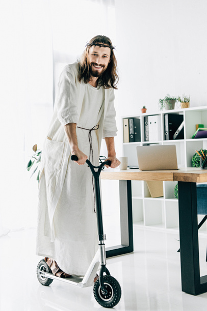 glücklicher Jesus in Dornenkrone und Robe auf Tretroller im modernen Büro  - Foto, Bild