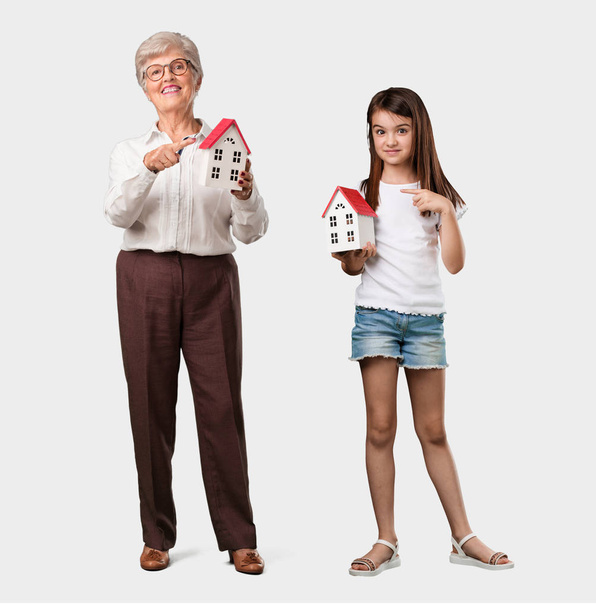 Полное тело пожилой женщины и ее внучки счастливой и уверенной в себе, показывающей миниатюрную модель дома, пытающейся ее продать, концепцию дома и семьи
 - Фото, изображение