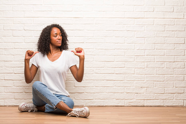 Giovane donna nera seduta su un pavimento di legno orgoglioso e fiducioso, puntando il dito contro muro di mattoni
 - Foto, immagini