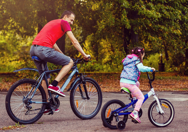 happy young father and his little daughter riding bikes in green park, side view - 写真・画像