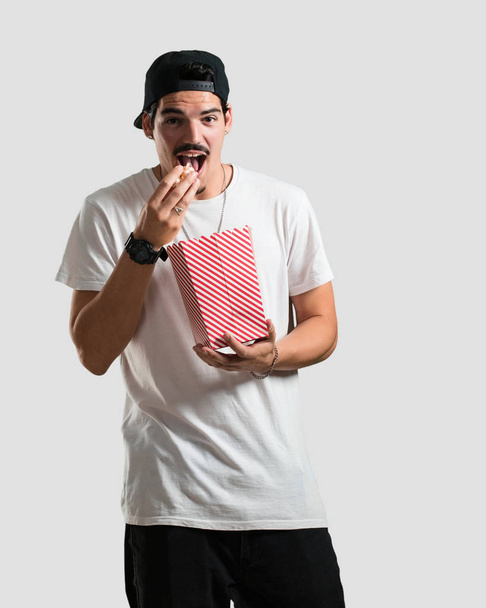 Jonge rapper man gelukkig en gefascineerd, verrast houden een gestreepte popcorn emmer, door de nieuwe film, ogen open en uiting van bewondering - Foto, afbeelding