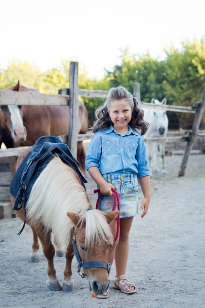 Маленькая улыбающаяся девочка с кудрявыми волосами, одетая в джинсы с пони в конюшне
 - Фото, изображение