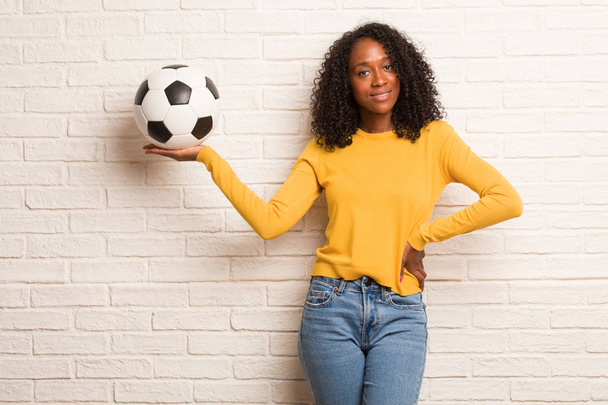 Młody czarny kobieta trzyma piłka nożna coś rękami przed mur z cegły - Zdjęcie, obraz