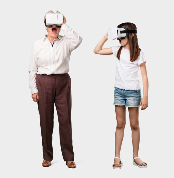 Ganzkörper einer älteren Dame und ihrer Enkelin aufgeregt und unterhaltsam, spielt mit Virtual-Reality-Brillen, erkundet eine Fantasiewelt, versucht etwas zu berühren - Foto, Bild