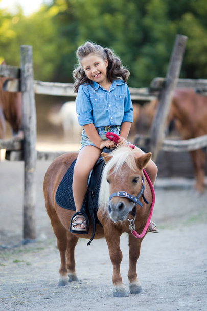 Une petite fille souriante aux cheveux bouclés habillée en jeans chevauchant un poney à l'écurie
 - Photo, image
