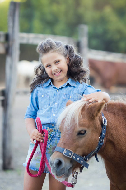 ein kleines lächelndes Mädchen mit lockigem Haar in Jeans mit einem Pony im Stall - Foto, Bild