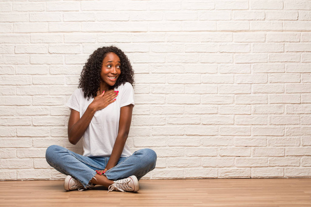 Young black woman sitting on a wooden floor doing a romantic gesture against brick wall - Foto, Bild