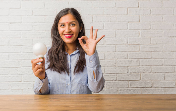 Young indian woman cheerful and confident doing ok gesture, excited and screaming, concept of approval and success. Holding a light bulb. - Foto, Bild