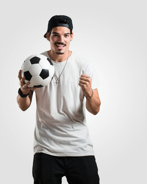 Νεαρός ράπερ άνθρωπος, χαμογελαστός και χαρούμενος, κρατώντας μια μπάλα ποδοσφαίρου, ανταγωνιστική στάση, ενθουσιασμένοι για να παίξετε ένα παιχνίδι - Φωτογραφία, εικόνα