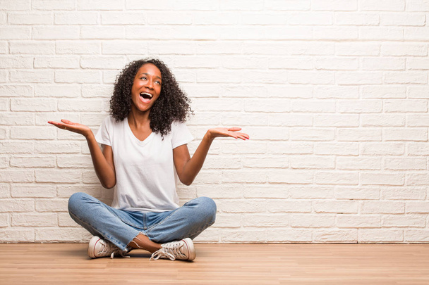 Joven mujer negra sentada en un piso de madera riendo y divirtiéndose contra la pared de ladrillo
 - Foto, imagen