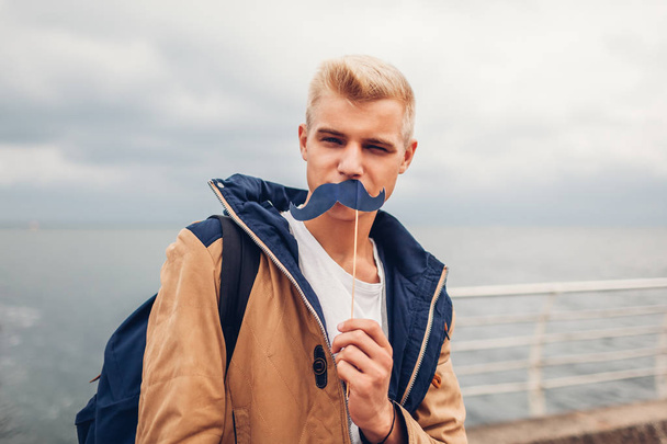 happy college student with backpack holding photo booth props moustache by sea. Guy having fun outdoors. Man winking - 写真・画像