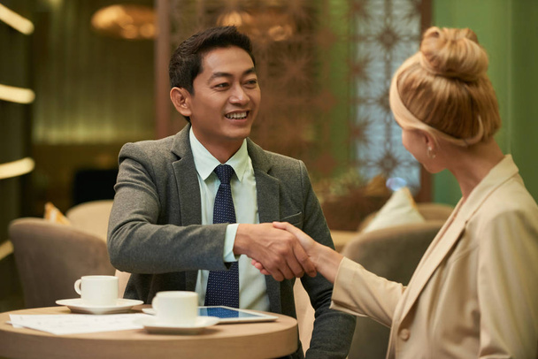 fröhlicher vietnamesischer Geschäftsmann schüttelt blonde Frau am Tisch im Café die Hand  - Foto, Bild
