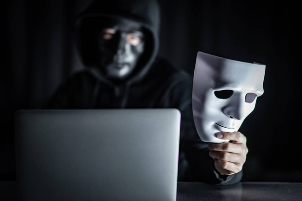 Záhadný muž s kapucí hacker černou maskou drží bílou maskou s přenosný počítač na stole. Anonymní sociální maskování. Ransomware cyber útoku nebo internetové koncepty zabezpečení - Fotografie, Obrázek