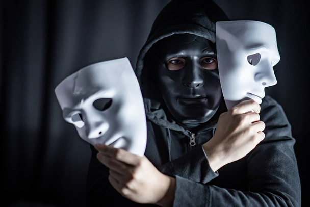 Tajemnica z kapturem człowiek sobie czarna maska, trzymając w ręku dwie białe maski. Anonimowe społecznej maskowania. Zaburzenia depresyjne lub choroby afektywnej dwubiegunowej. Koncepcja Halloween - Zdjęcie, obraz