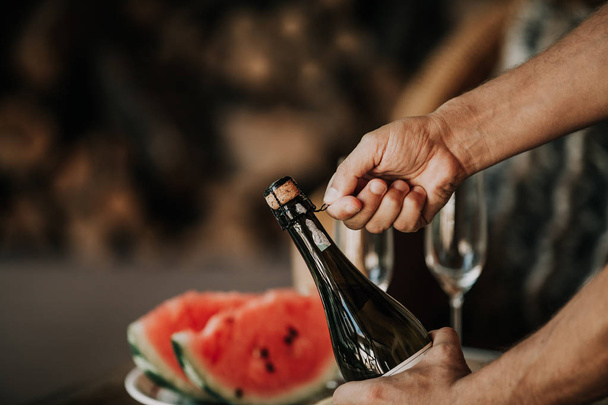 Hands twist the muse on a bottle of sparkling wine. Back on the glass table a plate with watermelon and frui - Foto, imagen