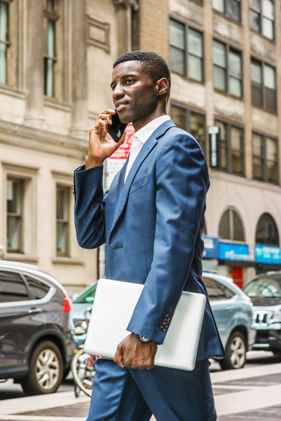 Jovem empresário afro-americano falando no celular, viajando, trabalhando em Nova York, vestindo terno azul, carregando computador portátil, andando na rua em Manhattan. Carros, edifícios em segundo plano - Foto, Imagem