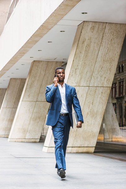 Uomo d'affari afroamericano che viaggia, lavora a New York, indossa un vestito blu, maglietta bianca, scarpe di pelle, porta computer portatile, cammina per strada fuori dall'edificio degli uffici, parla al telefono - Foto, immagini