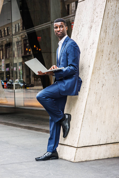 Giovane uomo d'affari afro-americano che lavora a New York, indossa vestito blu, camicia bianca, scarpe di pelle, in piedi contro la colonna sulla strada fuori dall'edificio degli uffici, tenendo il computer portatile, guardando altrove
 - Foto, immagini