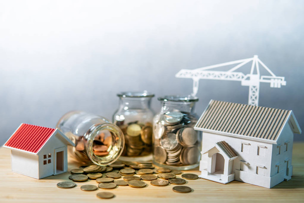 Haus- und Kranmodelle mit Münzen in Währungsgläsern auf einem Holztisch. Hypothekenzinsen für Eigenheime. Geld sparen. Immobilienentwicklung oder Immobilieninvestitionen. Geschäftskonzept der Bauindustrie - Foto, Bild