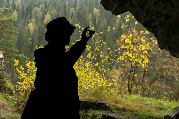 silueta de un geólogo examinando una muestra mineral encontrada en una cueva, sobre el fondo de una entrada con un bosque otoñal brillantemente iluminado
 - Foto, Imagen
