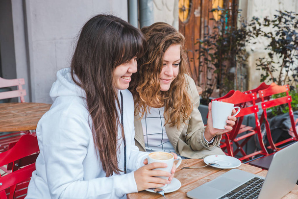 two adult young woman drinking coffee and looking at laptop in outdoors cafe. lifestyle concept - Foto, Imagem