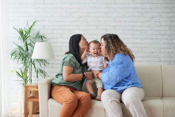 Multi-etnica coppia lesbica femminile con figlio a casa sul divano, donne che baciano le guance del bambino
   - Foto, immagini
