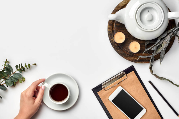 Genç kadın sağ üstünde çay bardağı, akıllı telefon ve su ısıtıcısı yanında beyaz masada boş defter sabah zamanında yazma - Fotoğraf, Görsel