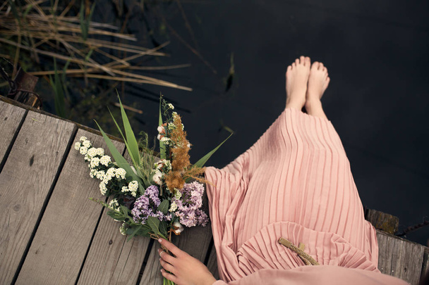 花の花束と農村の木造の橋に座っている桃色のドレスで美しい柔らかい少女. - 写真・画像