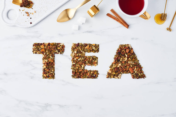Tea - word written by dried herbs and fruits on white marble background with tea utensils at the top. Flat lay with copy space. Tea concept, top view. - Foto, Imagen