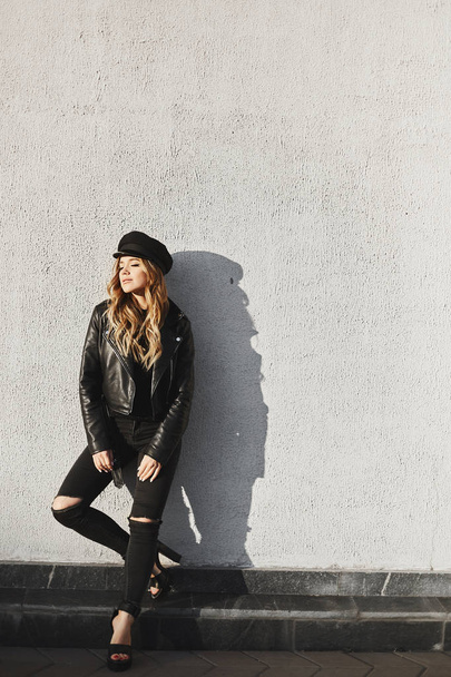 Fashionable and sensual blonde model girl in a black leather jacket, in black trendy jeans and in stylish hat smiling and posing with closed eyes outdoors - Foto, Bild