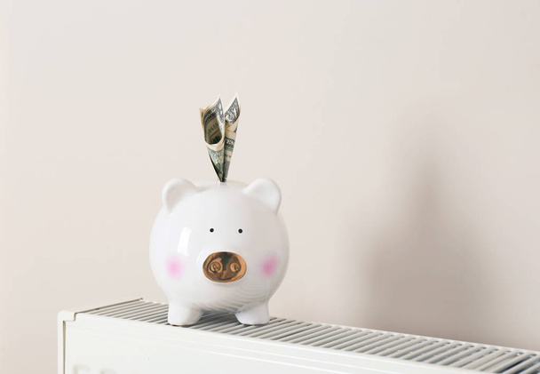 Piggy bank with money on heating radiator against light background - Foto, Imagen