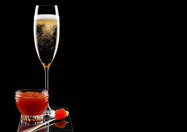 Elegant glass of yellow champagne with red caviar on golden spoon and glass container of caviar on marble board on black background - Foto, imagen