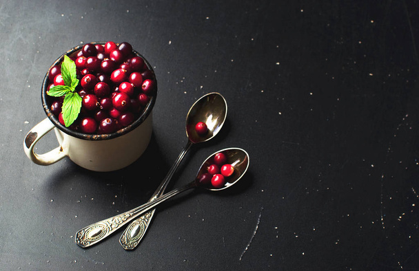 Fresh red cranberries with mint leaves in a white cup and spoons on a dark black background with copy space. Berry, holiday christmas background - Fotografie, Obrázek