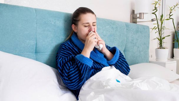 Portrait of sick young woman with flu lying in bed and using nasal spray - Fotó, kép