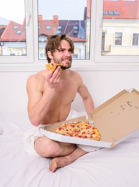 Break diet concept. Food delivery service. Man bearded handsome bachelor eating cheesy food for breakfast in bed. Man likes pizza for breakfast. Guy holds pizza box sit bed in bedroom or hotel room - Photo, Image