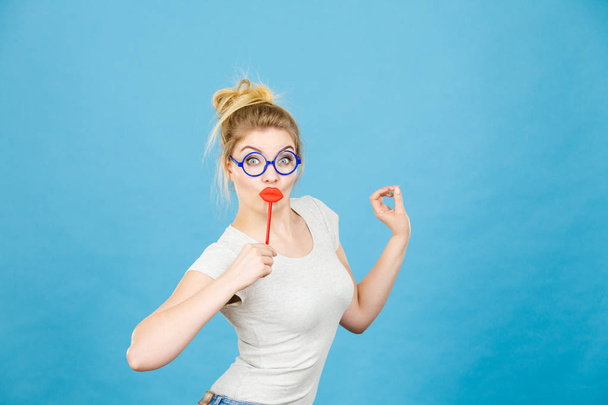 Crazy woman casual style nerdy glasses holding red fake lips on stick having fun, on blue. Photo take and carnival funny accessories concept. - Fotó, kép