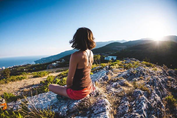 Uma menina está sentada no topo de uma montanha ao pôr-do-sol. Uma mulher está envolvida em ioga e senta-se em uma posição de lótus. A meditação está longe da civilização. Linda fêmea de volta. Vista das montanhas e do mar
. - Foto, Imagem