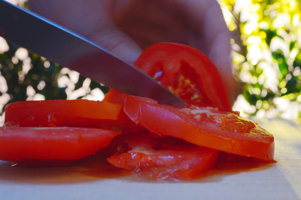 closeup slicing a tomato with a hand and knife on cutting board against the background of trees and sunset in the evening - Photo, Image