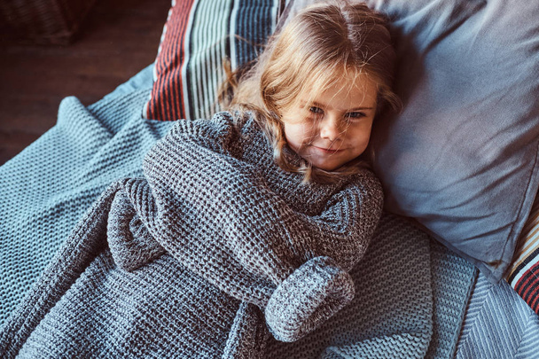 Close-up portrait of a little girl in warm sweater lying on bed. - Фото, изображение