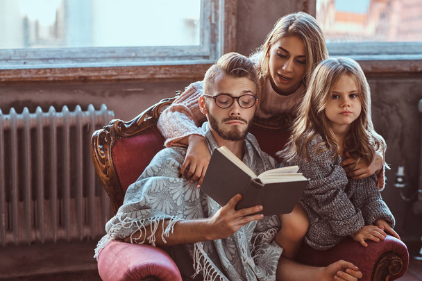 La unión familiar. Mamá, papá e hija leyendo un libro de cuentos juntos sentados en el sofá. Concepto de familia y paternidad
. - Foto, imagen
