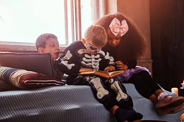 drei multirassische Kinder in gruseligen Kostümen, die Horrorgeschichten lesen, während sie in einem alten Haus auf dem Bett sitzen. - Foto, Bild